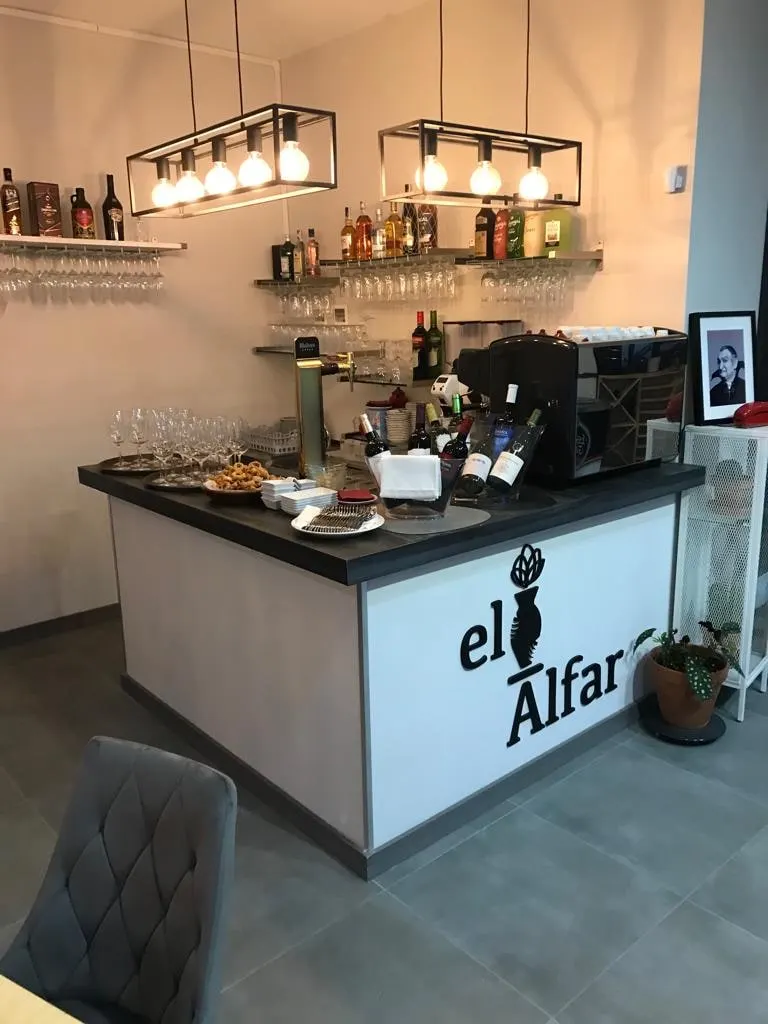 Barra de El Alfar con el logotipo del cántaro en el frente, botellas de vino y lámparas de bombillas encima