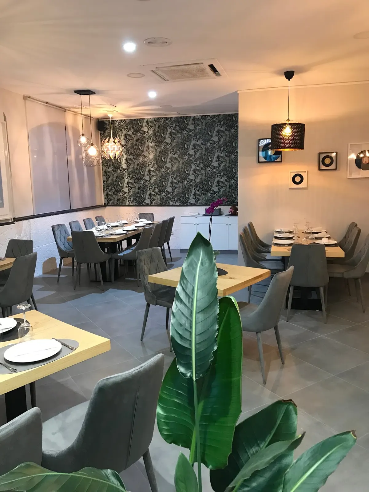 Comedor de El Alfar con mesas de madera clara, sillas de terciopelo gris, papel pintado de hojas y lámparas colgantes encendidas