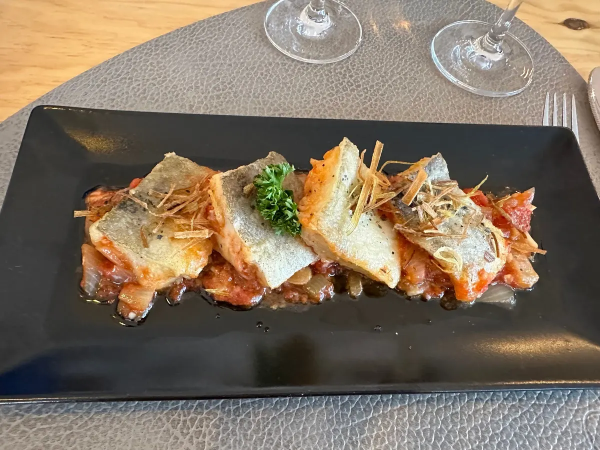 Lomos de pescado dorados sobre un sofrito de tomate, con puerro frito y perejil, en una fuente negra alargada