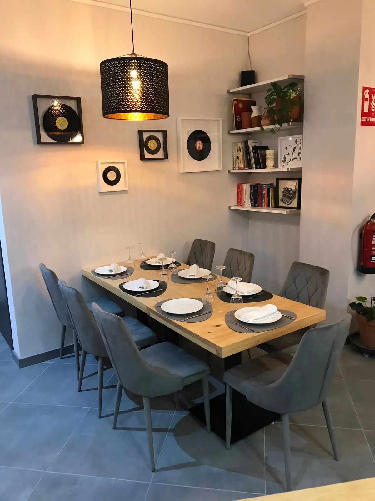 Mesa larga preparada junto a una pared con vinilos enmarcados, estantes con libros y una lámpara negra de luz cálida
